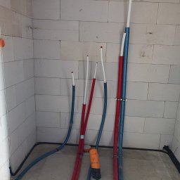 ETP Solution - Wnętrze w trakcie prac instalacyjnych: rury grzewcze (czerwone) i wodociągowe (niebieskie) biegnące wzdłuż ściany z bloczków, widoczna wiertarka Ryobi na podłodze.