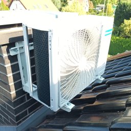 ETP Solution - Biała jednostka zewnętrzna klimatyzacji zamontowana na wspornikach do komina na dachu z ciemnej dachówki ceramicznej, widok z dołu.