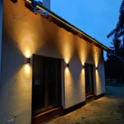 Elewacja domu oświetlona czterema podwójnymi lampami ściennymi, widoczne okna z brązowymi ramami, rynna i zbiornik na deszczówkę w tle, nocne ujęcie.