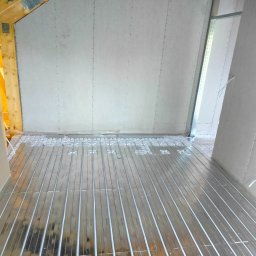 ETP Solution - Montaż ogrzewania podłogowego w pomieszczeniu z płytami gipsowo-kartonowymi na ścianach, widoczne rury grzewcze w metalowych profilach i elementy konstrukcji drewnianej dachu.