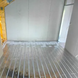 Montaż ogrzewania podłogowego w pomieszczeniu z płytami gipsowo-kartonowymi na ścianach, widoczne rury grzewcze w metalowych profilach i elementy konstrukcji drewnianej dachu.