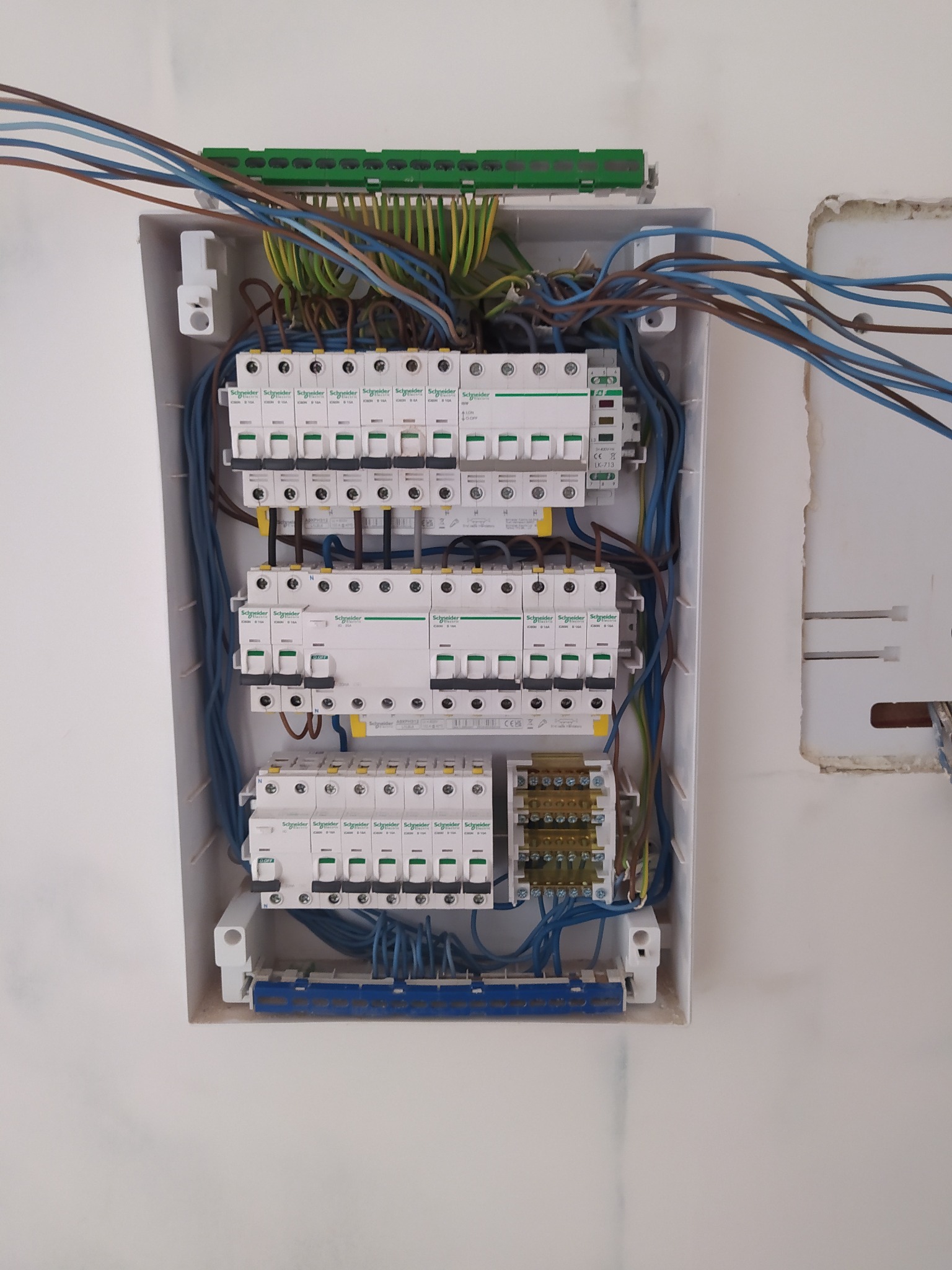 Otwarta skrzynka elektryczna z wieloma bezpiecznikami, przewodami w różnych kolorach i listwami zaciskowymi, zamontowana na białej ścianie.