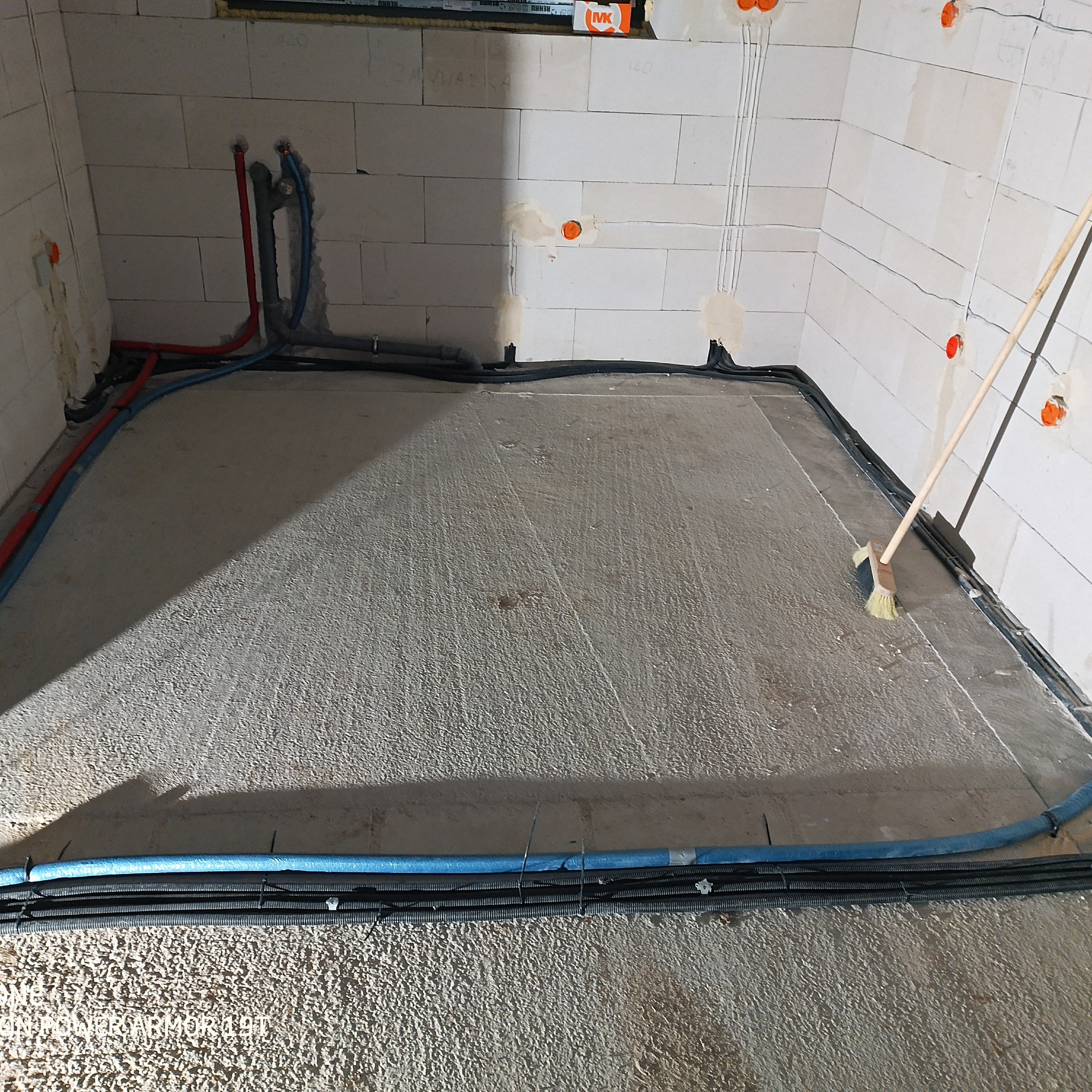 Wylewka betonowa z rozprowadzonymi rurami ogrzewania podłogowego i kablami elektrycznymi w surowym stanie, przygotowywana do dalszych prac wykończeniowych, z widoczną szczotką do zamiatania.