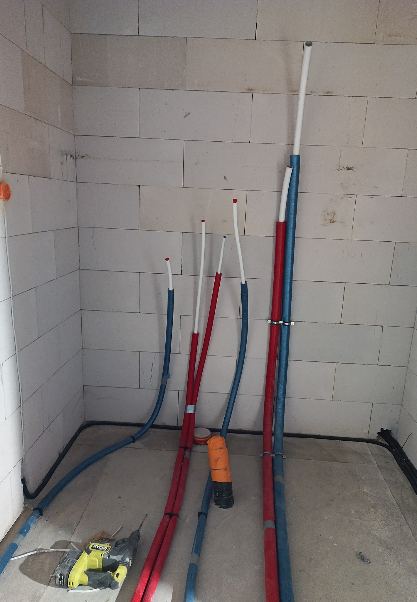 Wnętrze w trakcie prac instalacyjnych: rury grzewcze (czerwone) i wodociągowe (niebieskie) biegnące wzdłuż ściany z bloczków, widoczna wiertarka Ryobi na podłodze.