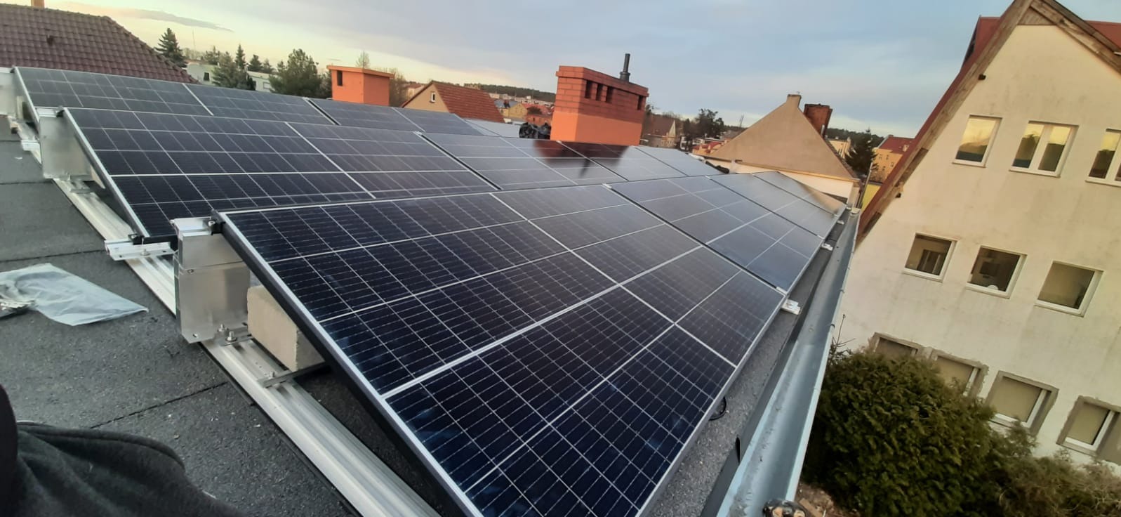 Panele słoneczne zamontowane na dachu budynku mieszkalnego, widoczne elementy montażowe i otoczenie miejskie z kominami i dachami innych budynków.