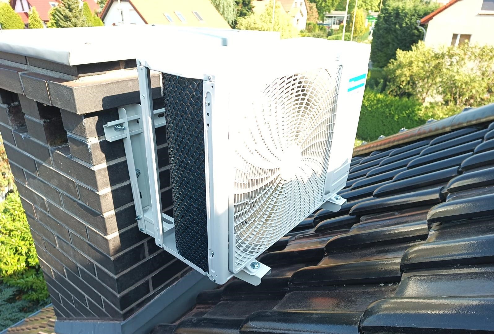 Biała jednostka zewnętrzna klimatyzacji zamontowana na wspornikach do komina na dachu z ciemnej dachówki ceramicznej, widok z dołu.