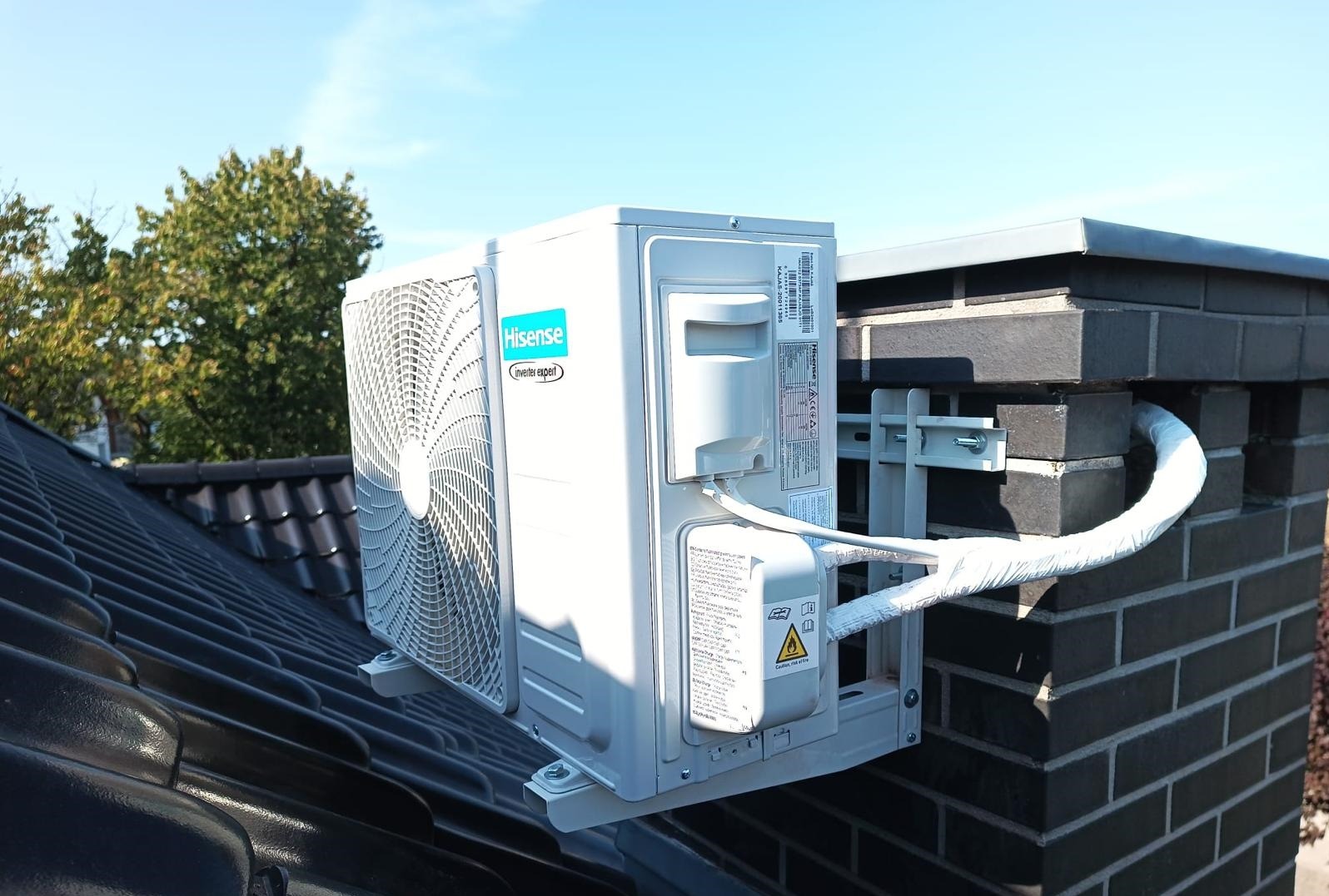 Zewnętrzna jednostka klimatyzacji Hisense zamontowana na wsporniku na dachu z czarnej dachówki, z widocznym przewodem w izolacji prowadzącym do ściany z cegły.