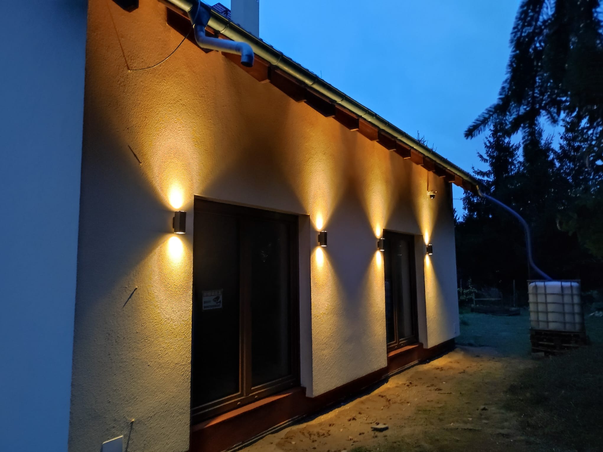 Elewacja domu oświetlona czterema podwójnymi lampami ściennymi, widoczne okna z brązowymi ramami, rynna i zbiornik na deszczówkę w tle, nocne ujęcie.