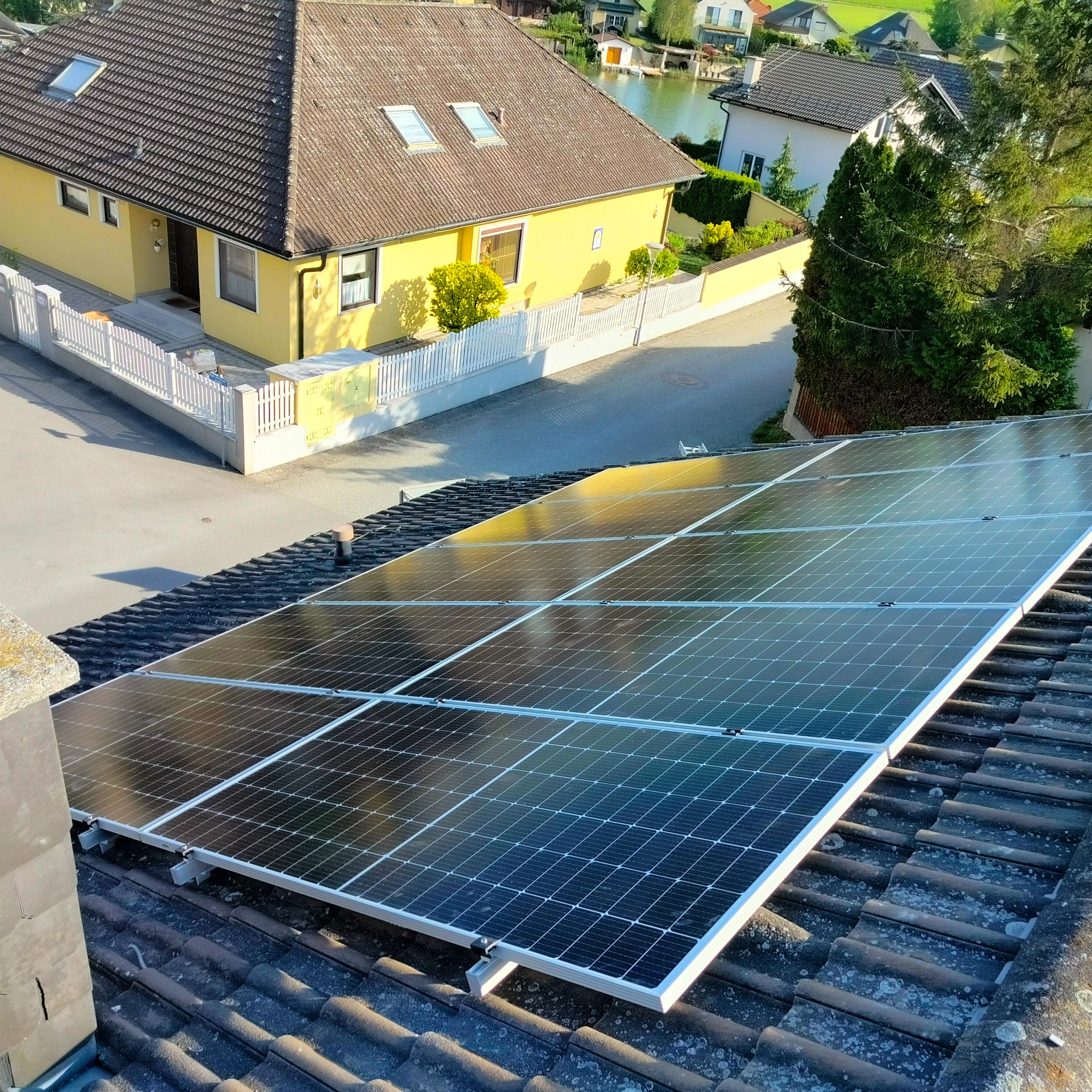 Panele słoneczne na brązowym, lekko zabrudzonym dachu domu z widokiem na okolicę z jeziorem w tle.