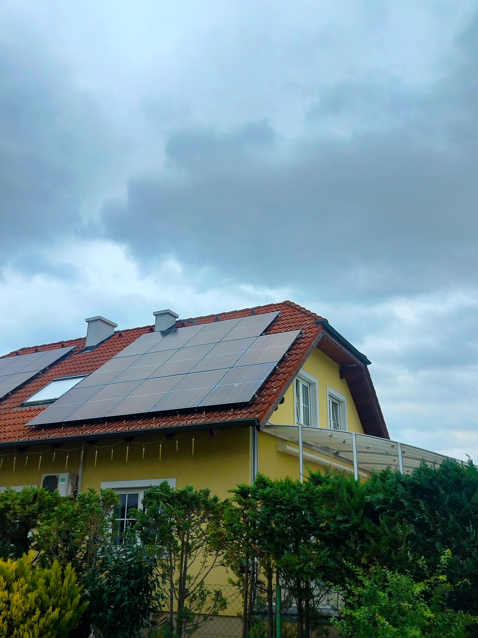 Żółty dom jednorodzinny z panelami fotowoltaicznymi na czerwonym dachu, widoczne dwa kominy i okno dachowe, tło pochmurne niebo.