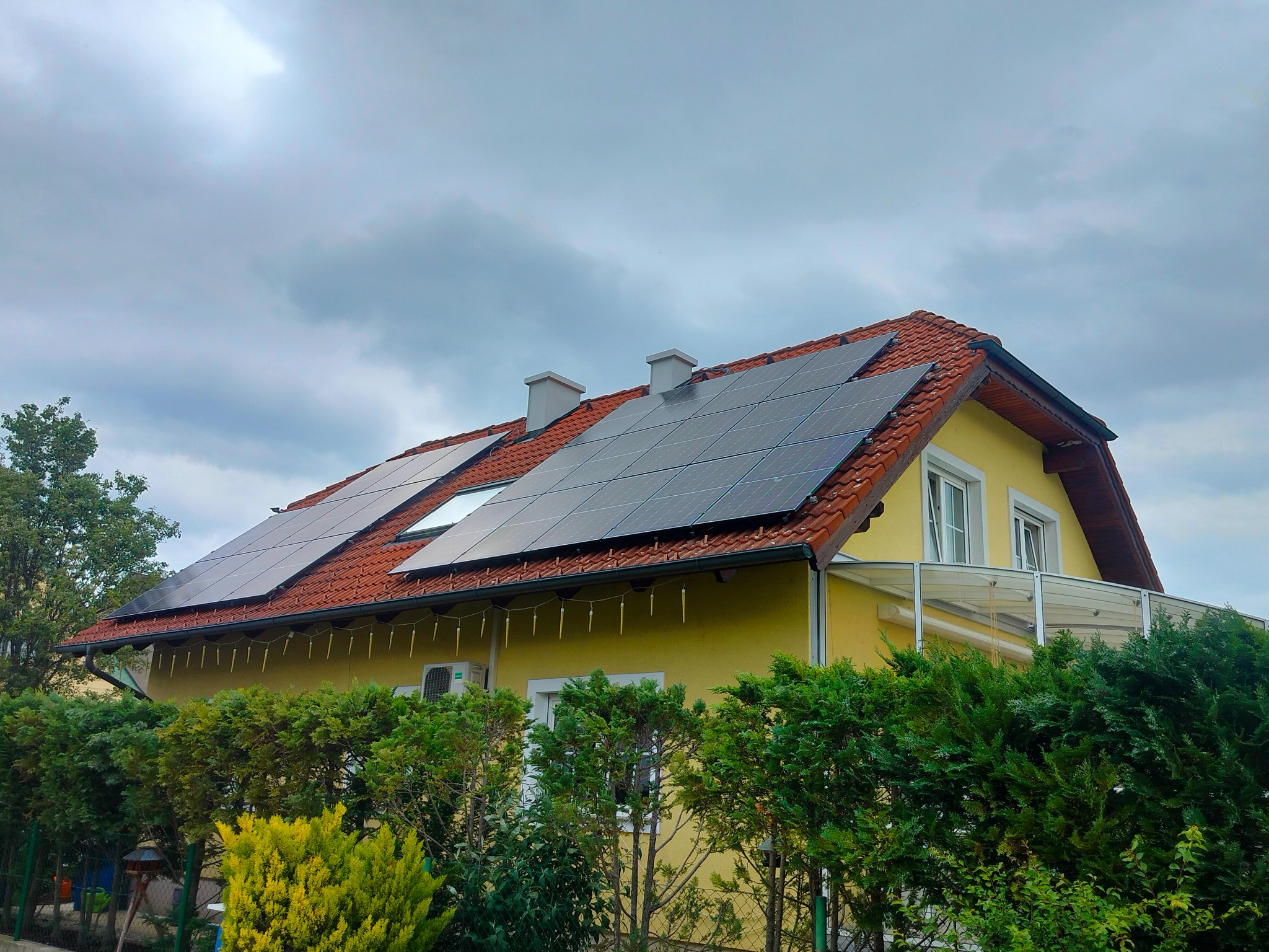 Żółty dom jednorodzinny z pomarańczową dachówką, na której zamontowano panele fotowoltaiczne, widoczne również dwa kominy i balkon z przezroczystą balustradą, a w tle pochmurne niebo.