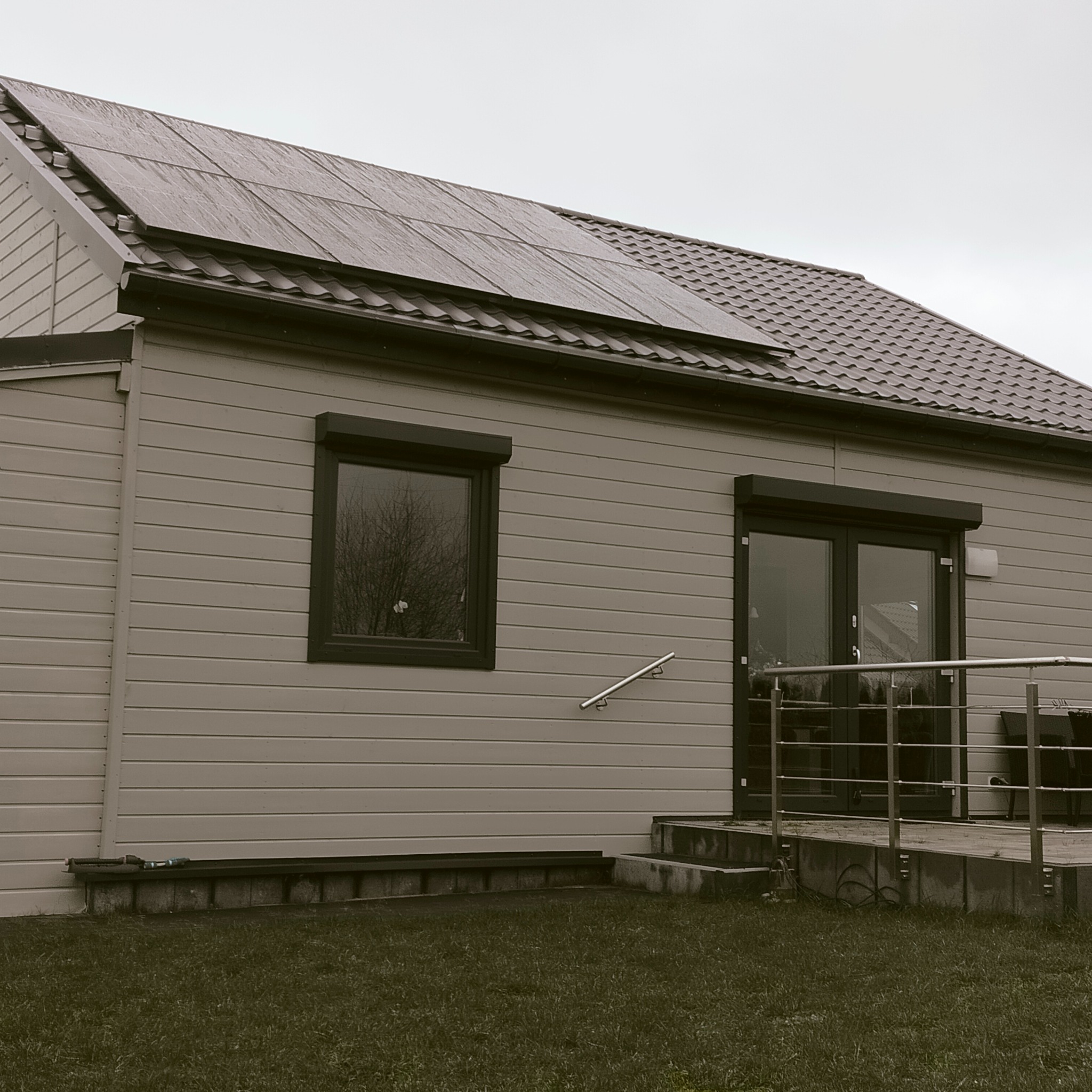 Dom jednorodzinny z panelami fotowoltaicznymi na dachu, szara elewacja z oknem i drzwiami balkonowymi, trawnik przed budynkiem, pochmurne niebo.