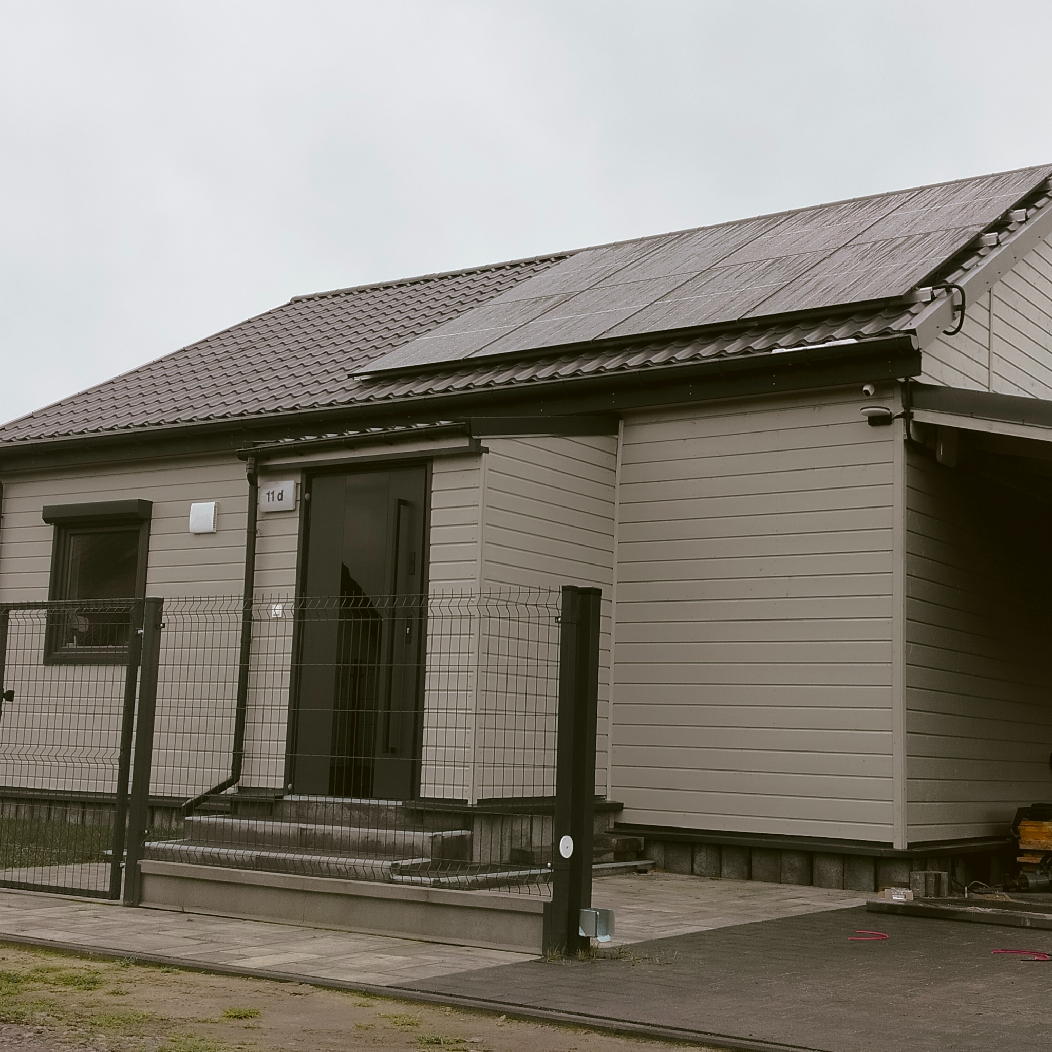 Dom jednorodzinny z panelami słonecznymi na dachu pokrytym brązową dachówką, ogrodzony siatką, w pochmurny dzień.