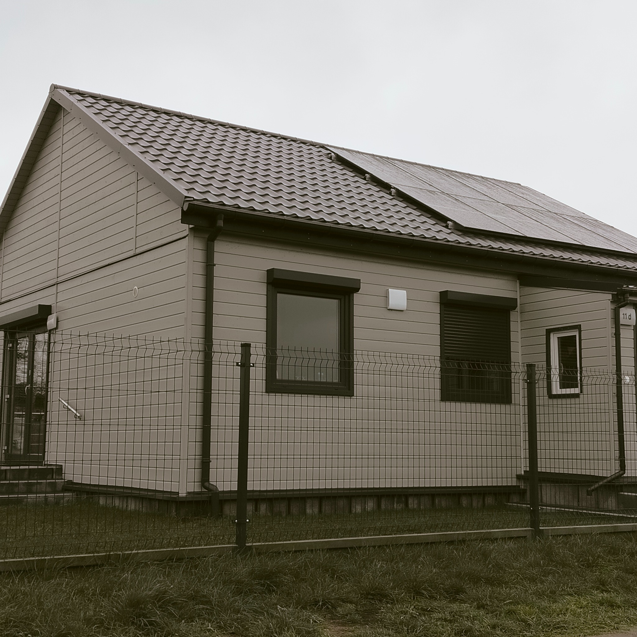 Dom jednorodzinny z panelami fotowoltaicznymi na dachu, widok zza ogrodzenia z siatki, szare kolory, pochmurne niebo.