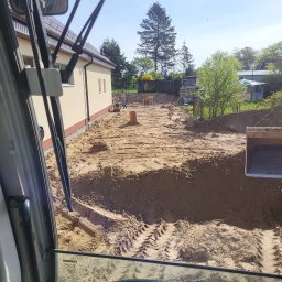 Prace ziemne - Widok z kabiny koparki na plac budowy z wykopem wzdłuż fundamentów budynku, widoczne ślady opon i pracujący robotnicy w oddali.