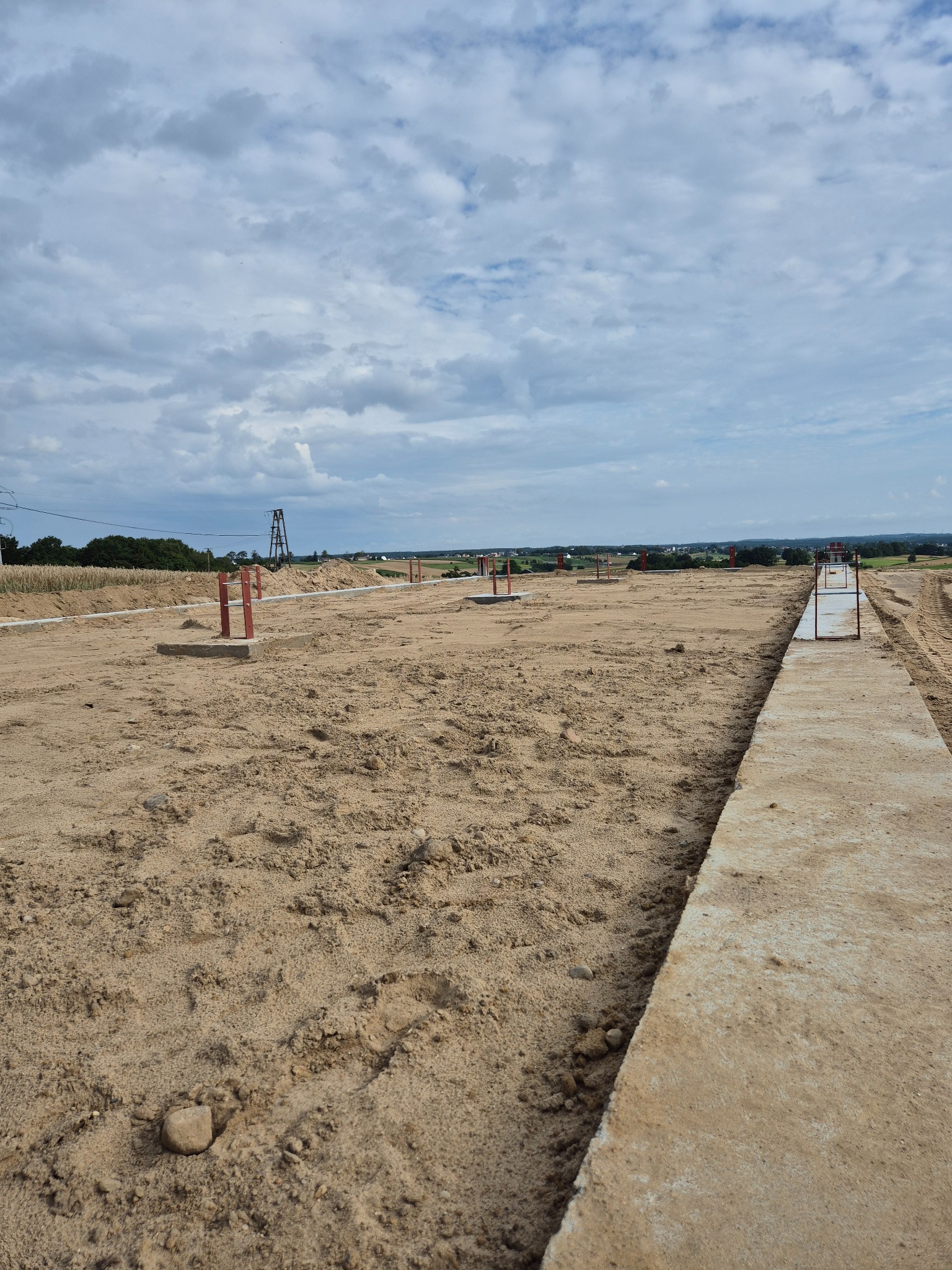 Przygotowany teren pod budowę z wytyczonymi fundamentami, widoczne betonowe ławy fundamentowe i metalowe znaczniki, tło stanowią pola i pochmurne niebo.