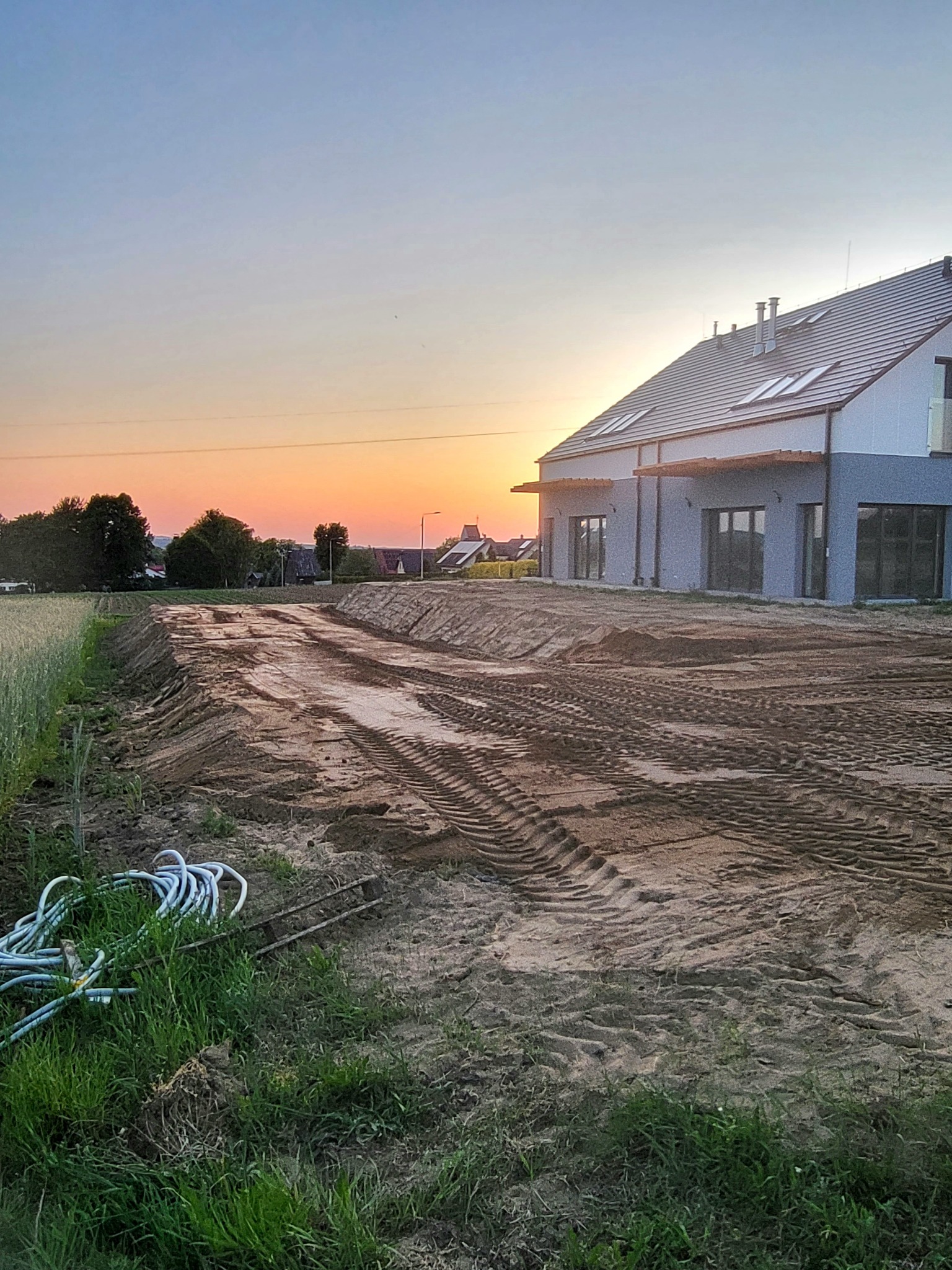 Przygotowany teren pod budowę domu jednorodzinnego, widoczne ślady opon maszyn budowlanych na wyrównanej powierzchni ziemi, w tle zachodzące słońce.