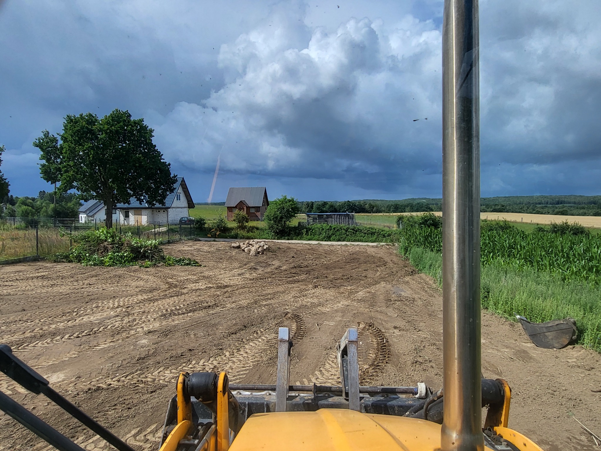 Widok z kabiny traktora na wyrównany teren w Luzinie, z widocznymi śladami opon i przygotowanym podłożem pod budowę, w tle zabudowania gospodarcze i burzowe chmury.