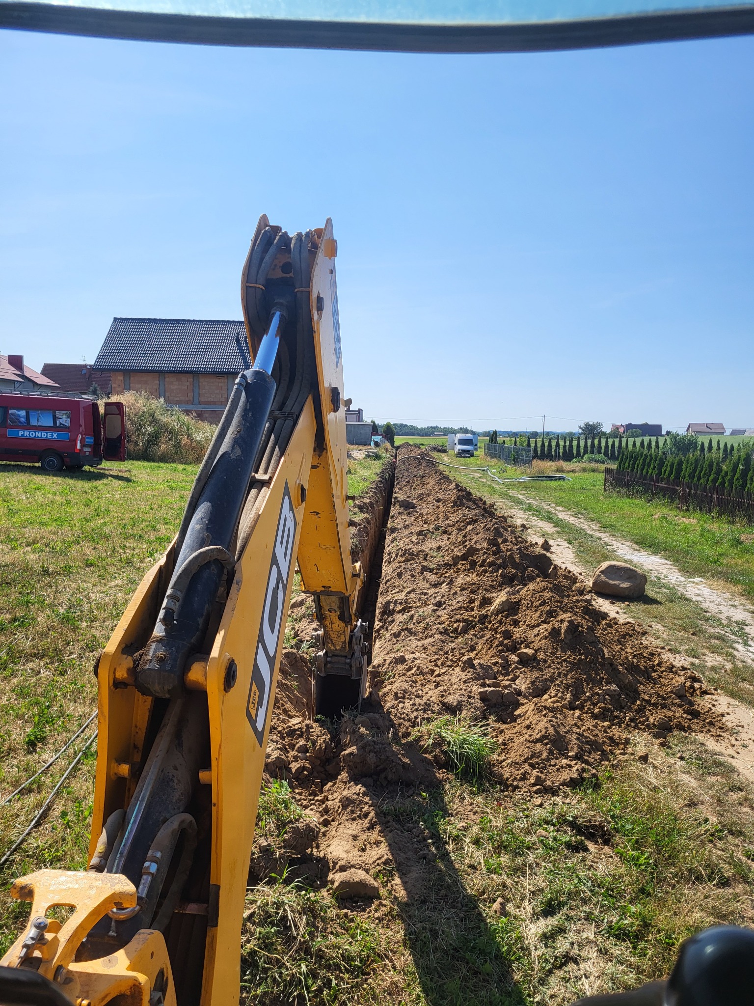 Żółta koparka JCB w trakcie wykonywania wykopu w gruncie, widok z kabiny operatora, w tle zabudowa jednorodzinna i błękitne niebo.