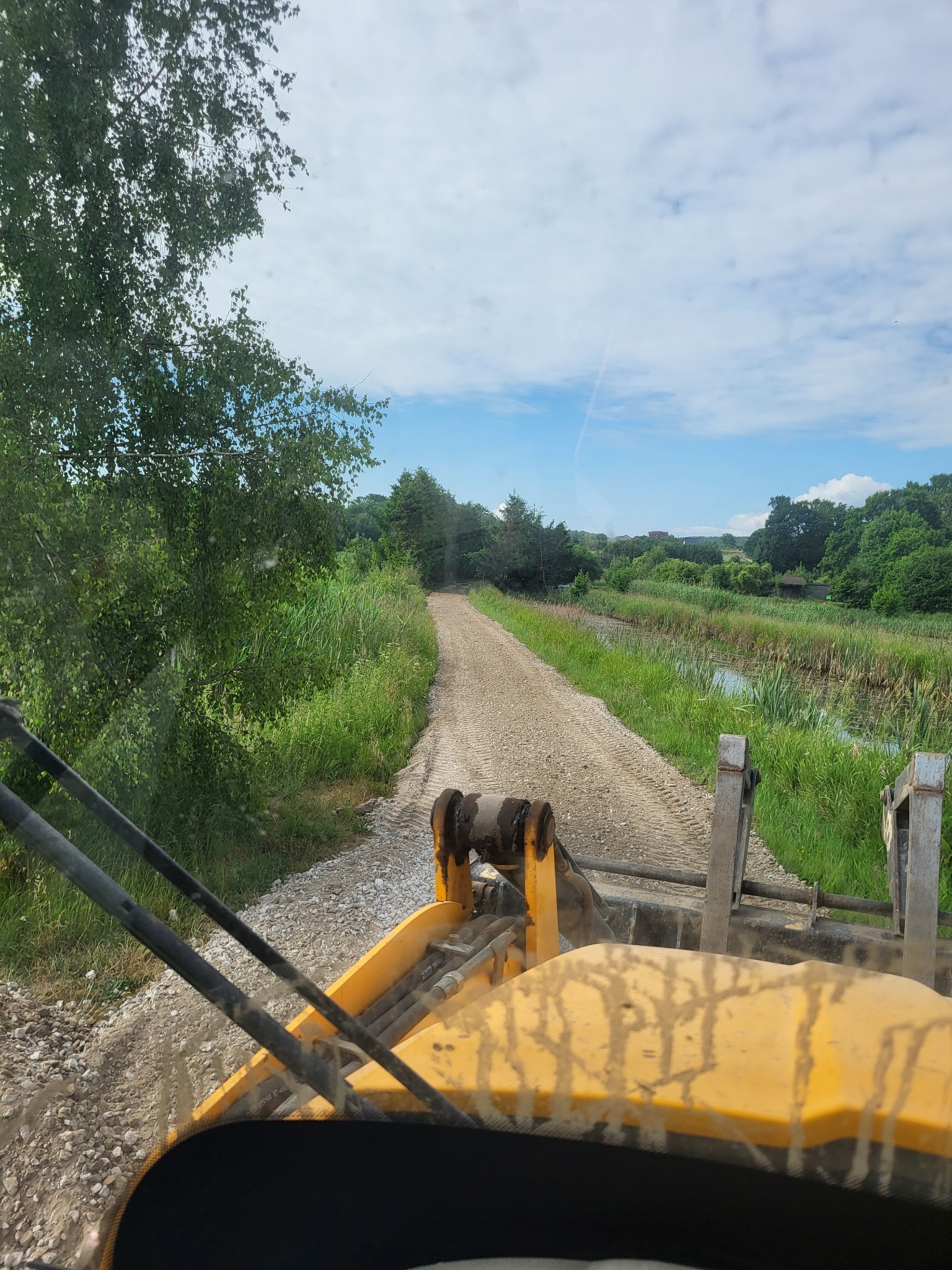 Widok z kabiny żółtej maszyny budowlanej na utwardzaną żwirową drogę, w tle zielona łąka i drzewa pod błękitnym, pochmurnym niebem.