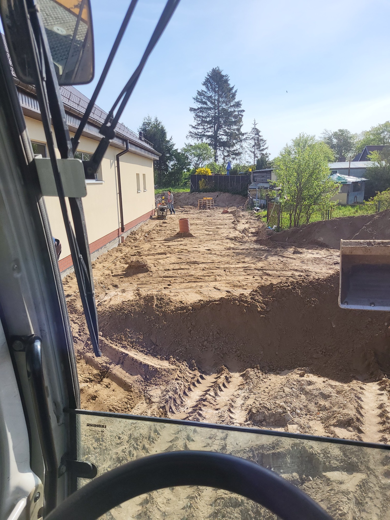 Widok z kabiny koparki na plac budowy z wykopem wzdłuż fundamentów budynku, widoczne ślady opon i pracujący robotnicy w oddali.