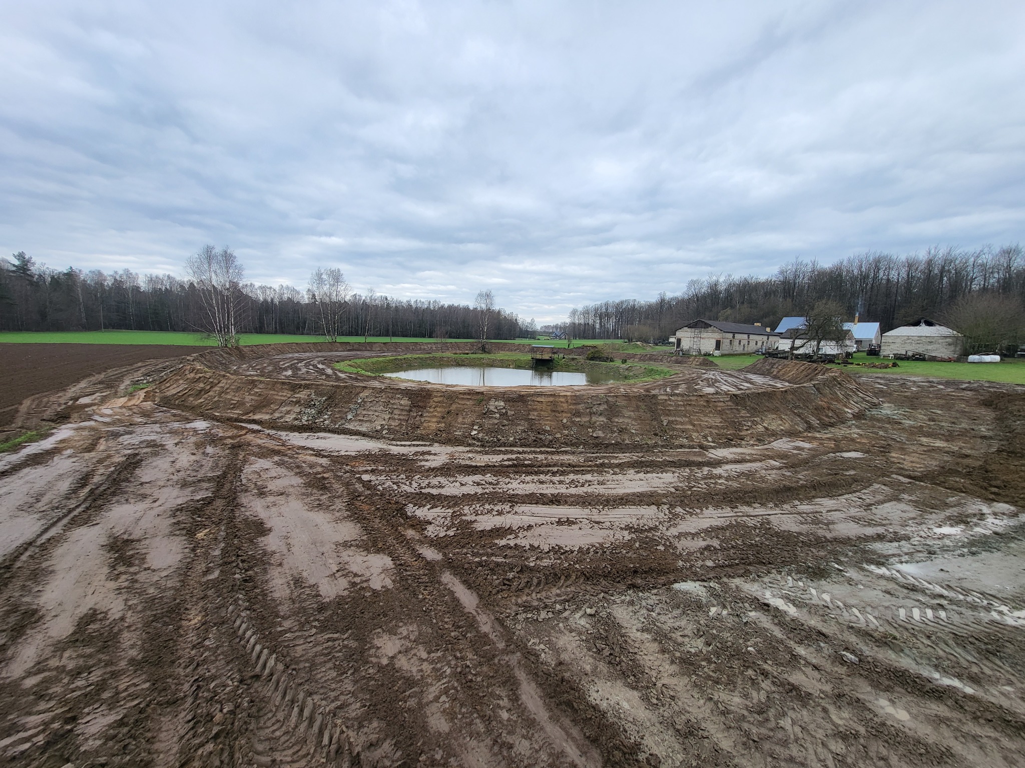 Szeroki kadr na okrągły zbiornik wodny w trakcie budowy, otoczony ziemią z wyraźnymi śladami opon, w tle budynki gospodarcze i las pod pochmurnym niebem.