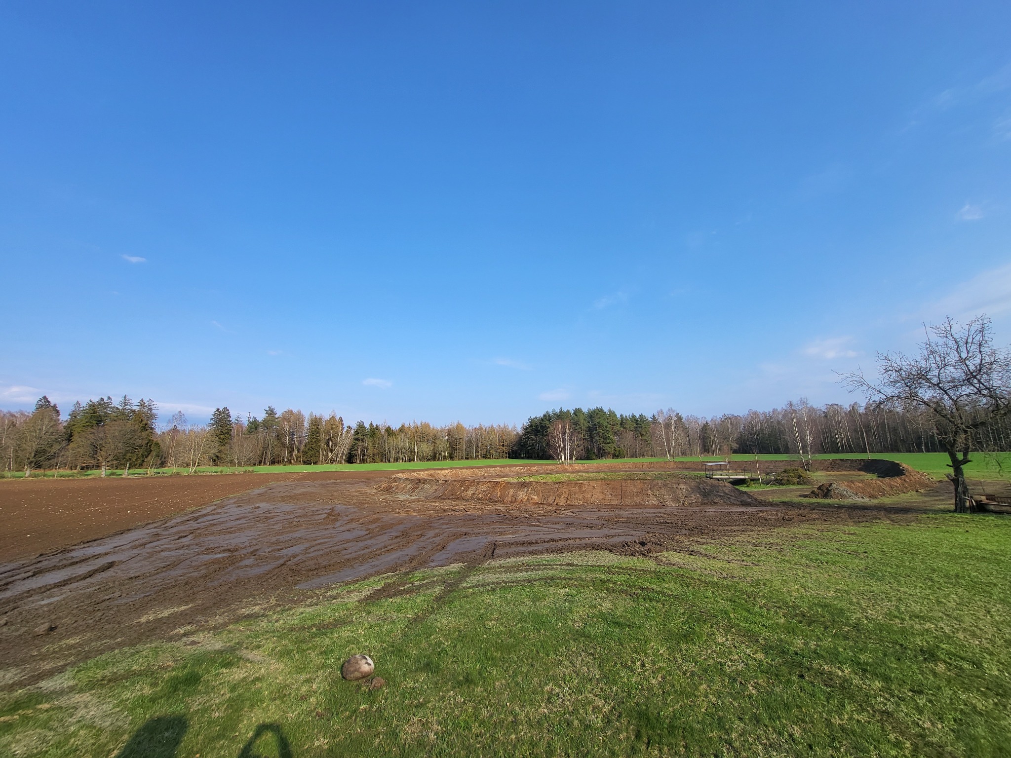 Świeżo wyrównany teren pod budowę, widoczne ślady opon w błocie, w tle las mieszany i błękitne niebo.