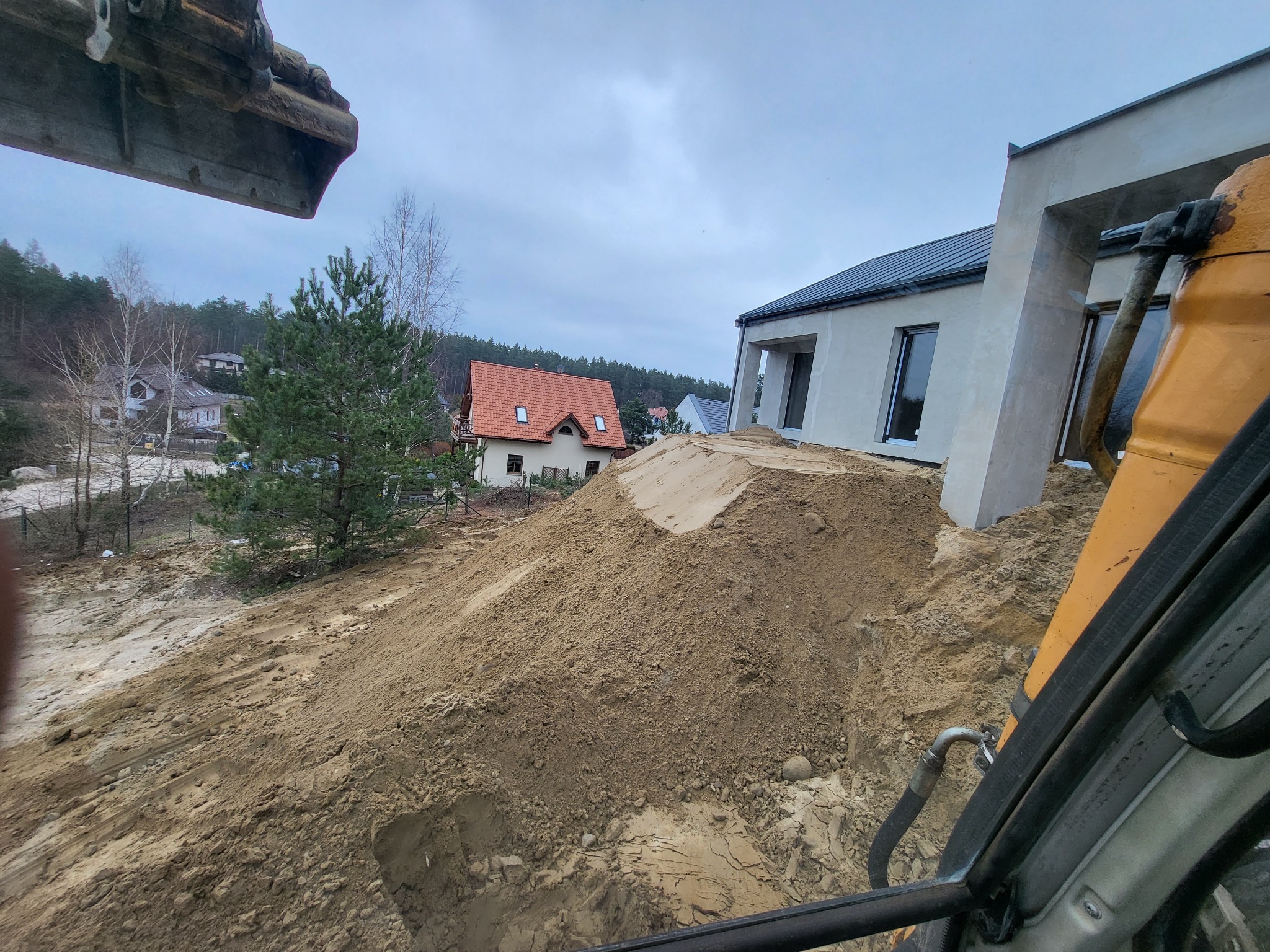 Żółta koparka przygotowuje teren pod budowę nowoczesnego domu; widoczna duża pryzma piasku i fragmenty lasu w tle.