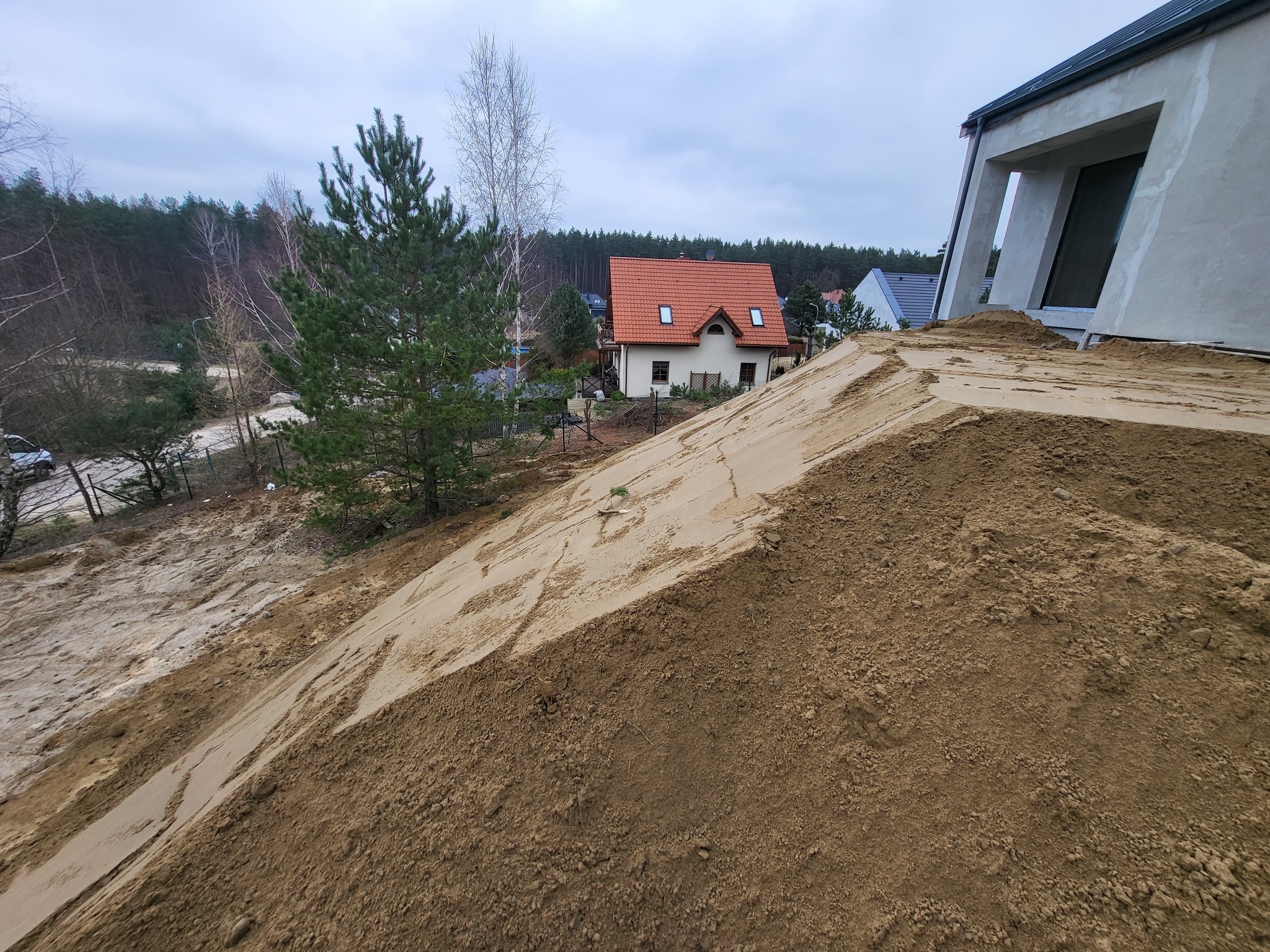 Kupa piasku usypana przy nowoczesnym domu w trakcie budowy, widok na okoliczną zabudowę i las w tle.