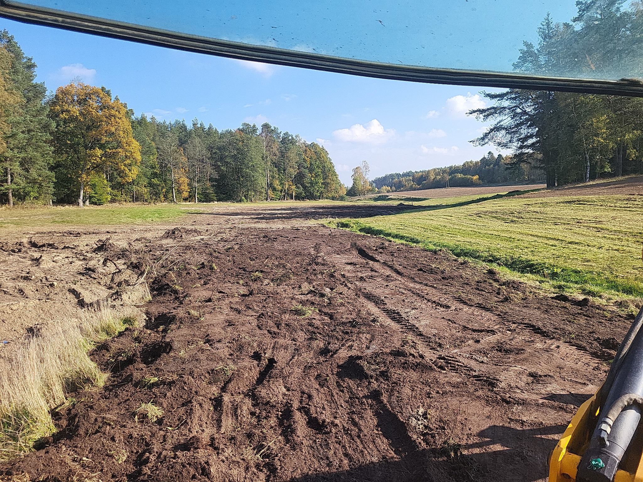 Widok z kabiny maszyny budowlanej na wyrównany teren, z widocznymi śladami opon, w tle las i błękitne niebo.
