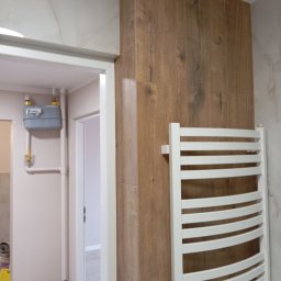 Usługi Remontowe Krzysztof Maciążka - Wykończona łazienka z płytkami imitującymi drewno na ścianie, biały grzejnik drabinkowy i widoczny licznik gazu w tle.