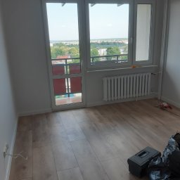 Usługi Remontowe Krzysztof Maciążka - Pokój po remoncie z drewnianą podłogą, białymi ścianami, grzejnikiem i widokiem z okna na miasto, widoczna skrzynka z narzędziami i worki na śmieci.