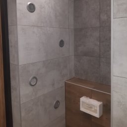 Usługi Remontowe Krzysztof Maciążka - Wykończona łazienka z szarymi płytkami imitującymi beton, drewnianym obudowaniem wanny i kratkami wentylacyjnymi w ścianie, widok na narożnik pomieszczenia.