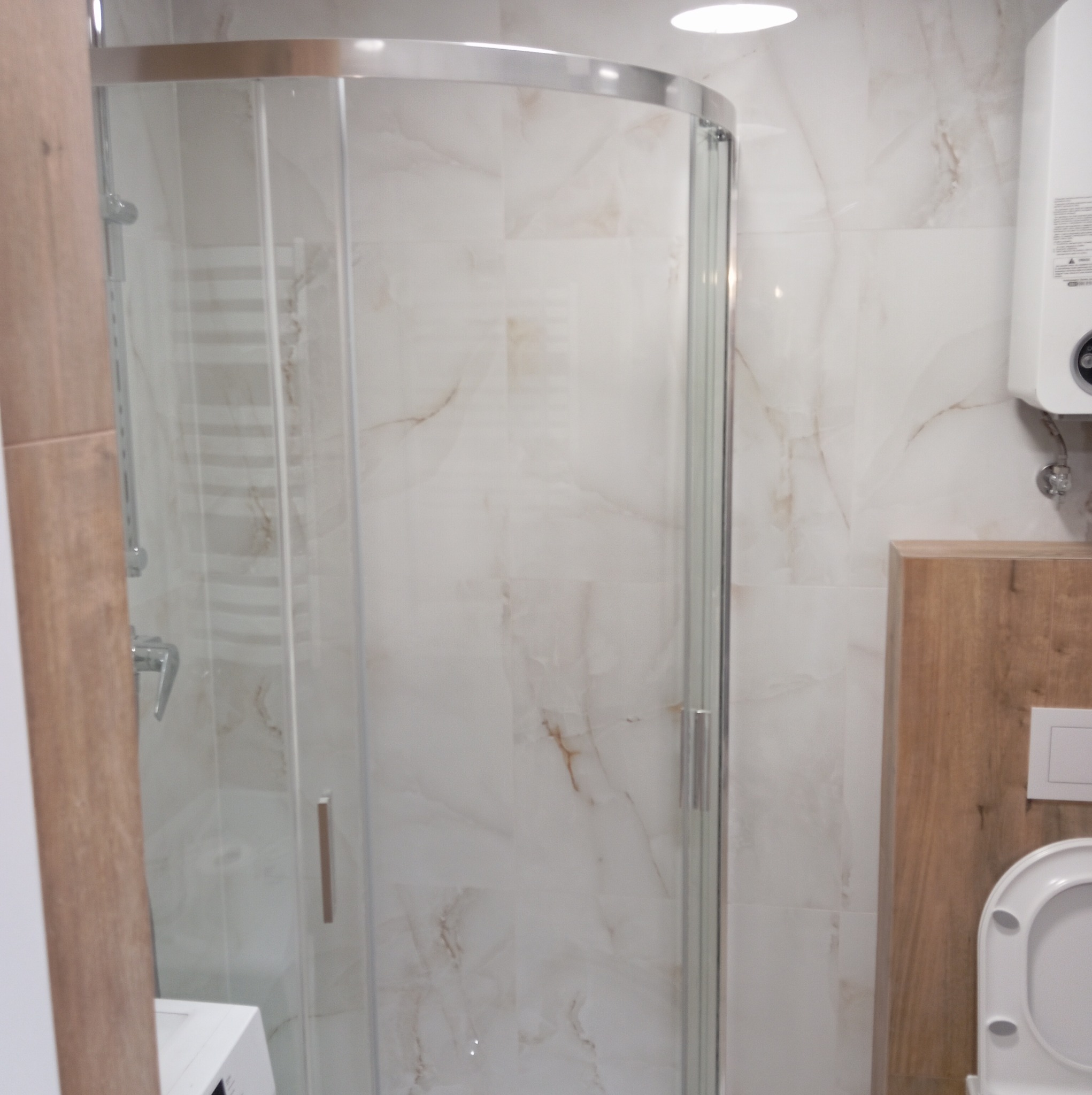 Wykończona łazienka: kabina prysznicowa z chromowanymi profilami, marmurowe płytki w odcieniach bieli i beżu, podwieszana toaleta z drewnianą obudową, bojler elektryczny na ścianie.