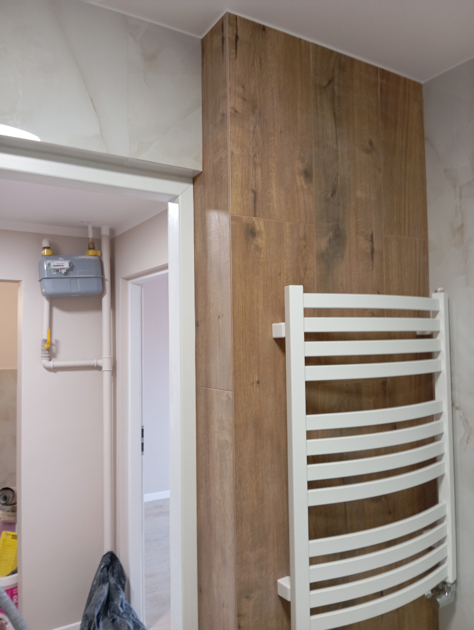 Wykończona łazienka z płytkami imitującymi drewno na ścianie, biały grzejnik drabinkowy i widoczny licznik gazu w tle.