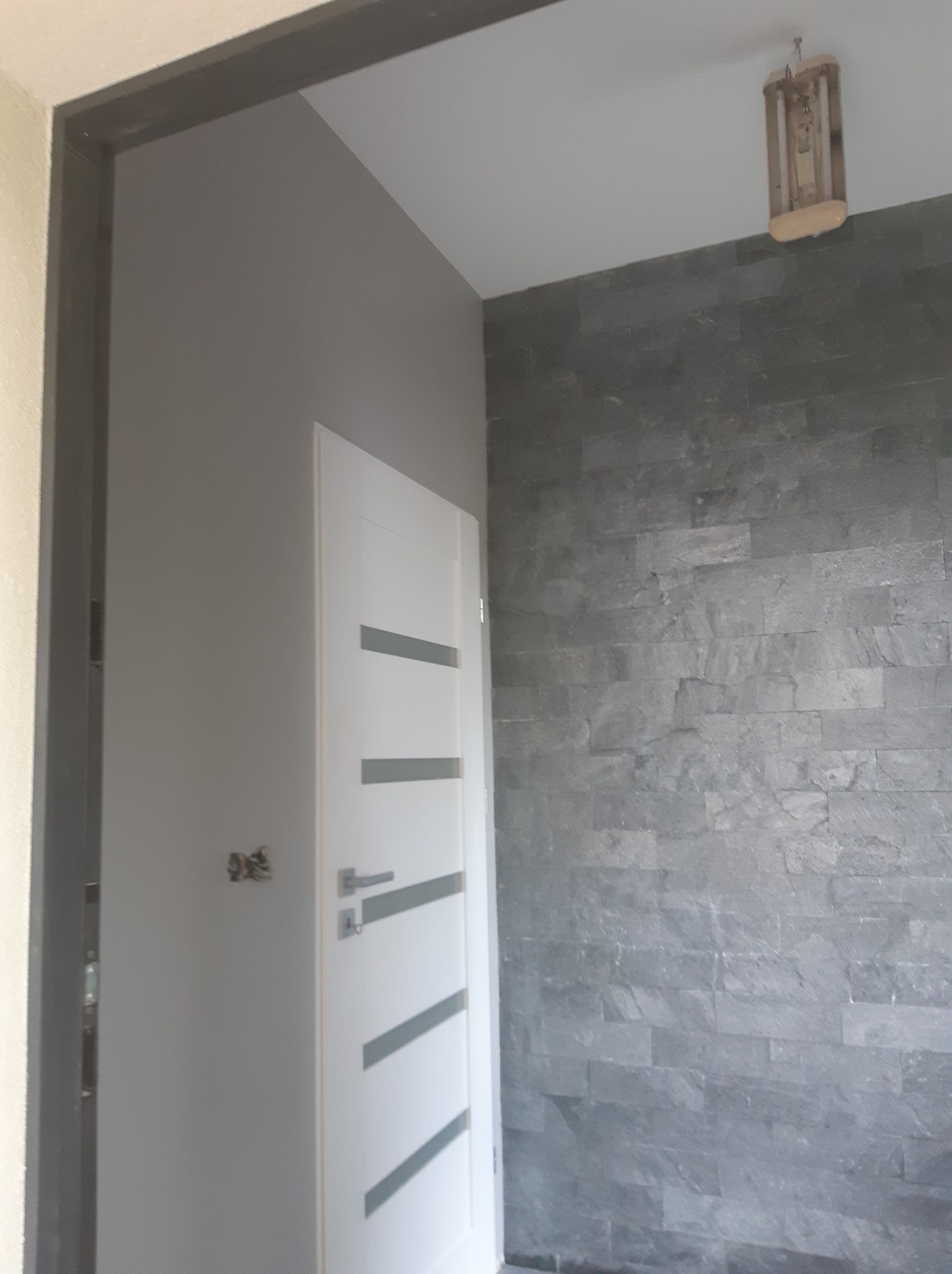 Szara ściana wykończona kamieniem dekoracyjnym, białe drzwi z poziomymi, szarymi paskami, widoczna ościeżnica i lampa sufitowa z drewnianą obudową.