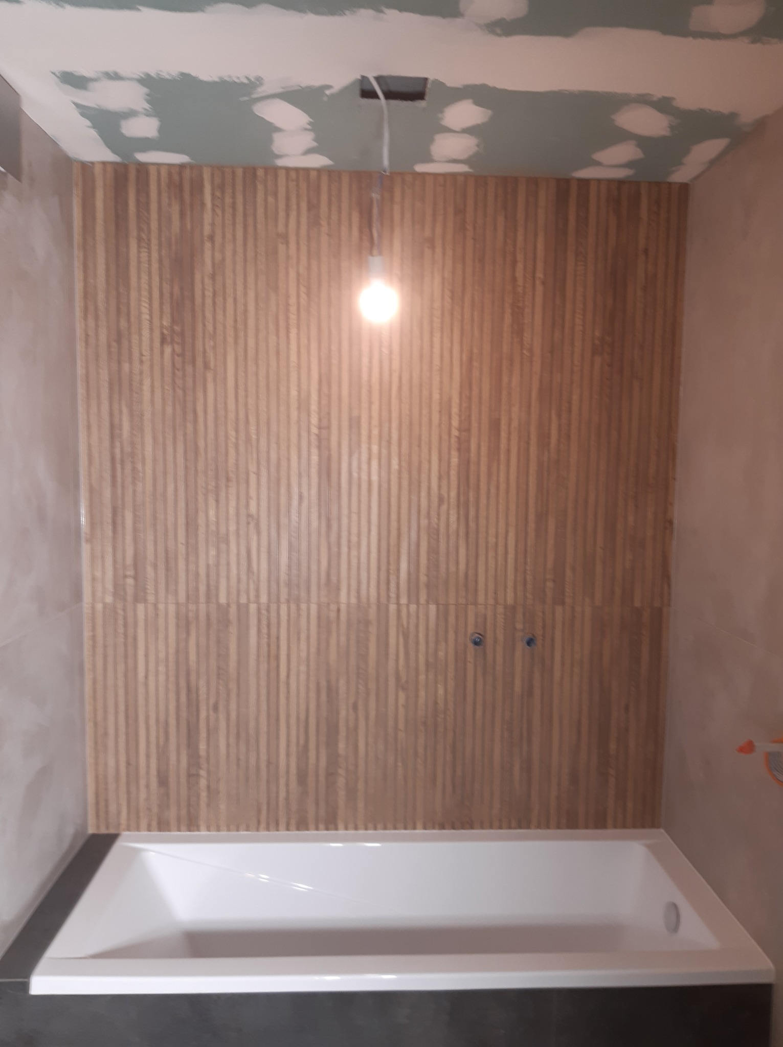 Wnętrze łazienki w trakcie prac wykończeniowych: ściana wyłożona panelami drewnopodobnymi, biała wanna, sufit z widocznymi śladami szpachlowania i przygotowania pod malowanie, wisząca żarówka bez...