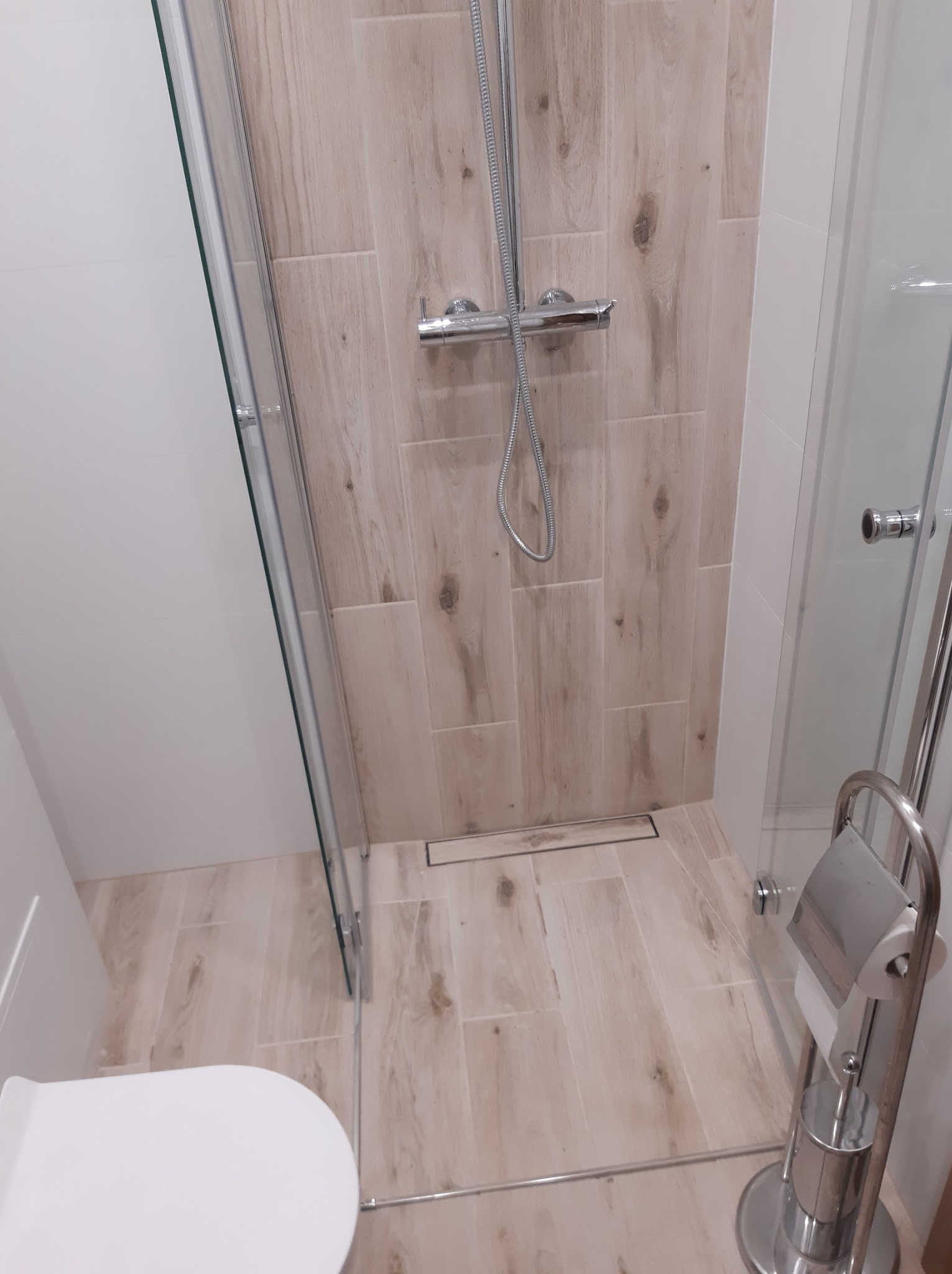 Nowoczesna kabina prysznicowa z odpływem liniowym, wykończona płytkami imitującymi drewno, z widocznym fragmentem toalety i chromowanym uchwytem na papier toaletowy.