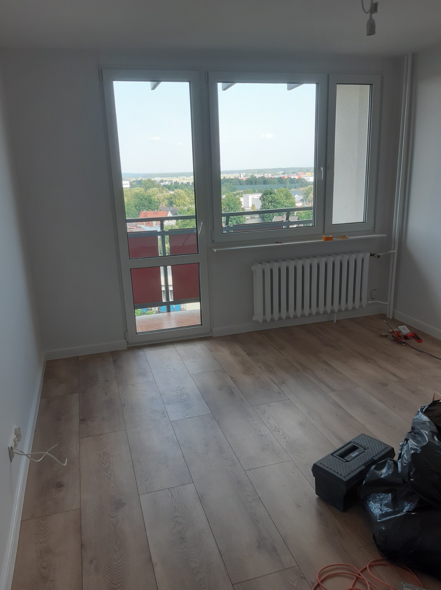 Pokój po remoncie z drewnianą podłogą, białymi ścianami, grzejnikiem i widokiem z okna na miasto, widoczna skrzynka z narzędziami i worki na śmieci.