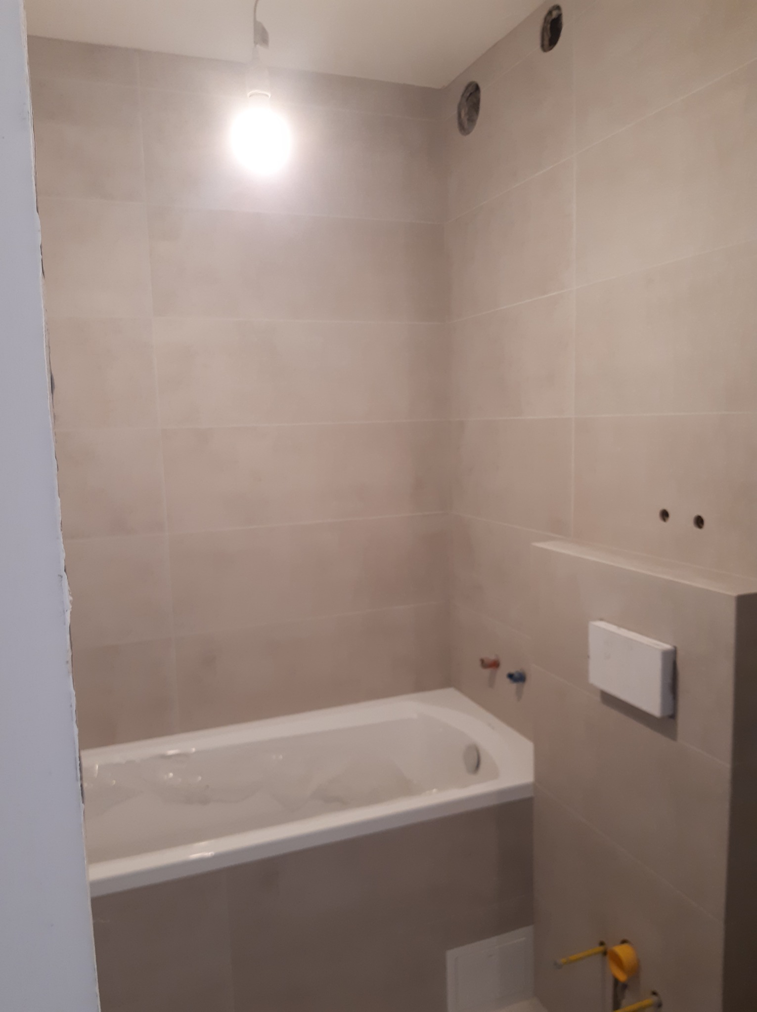 Beżowa łazienka z wanną, widoczne przyłącza hydrauliczne i otwory w ścianie pod oświetlenie, elementy instalacji sanitarnej w trakcie montażu.