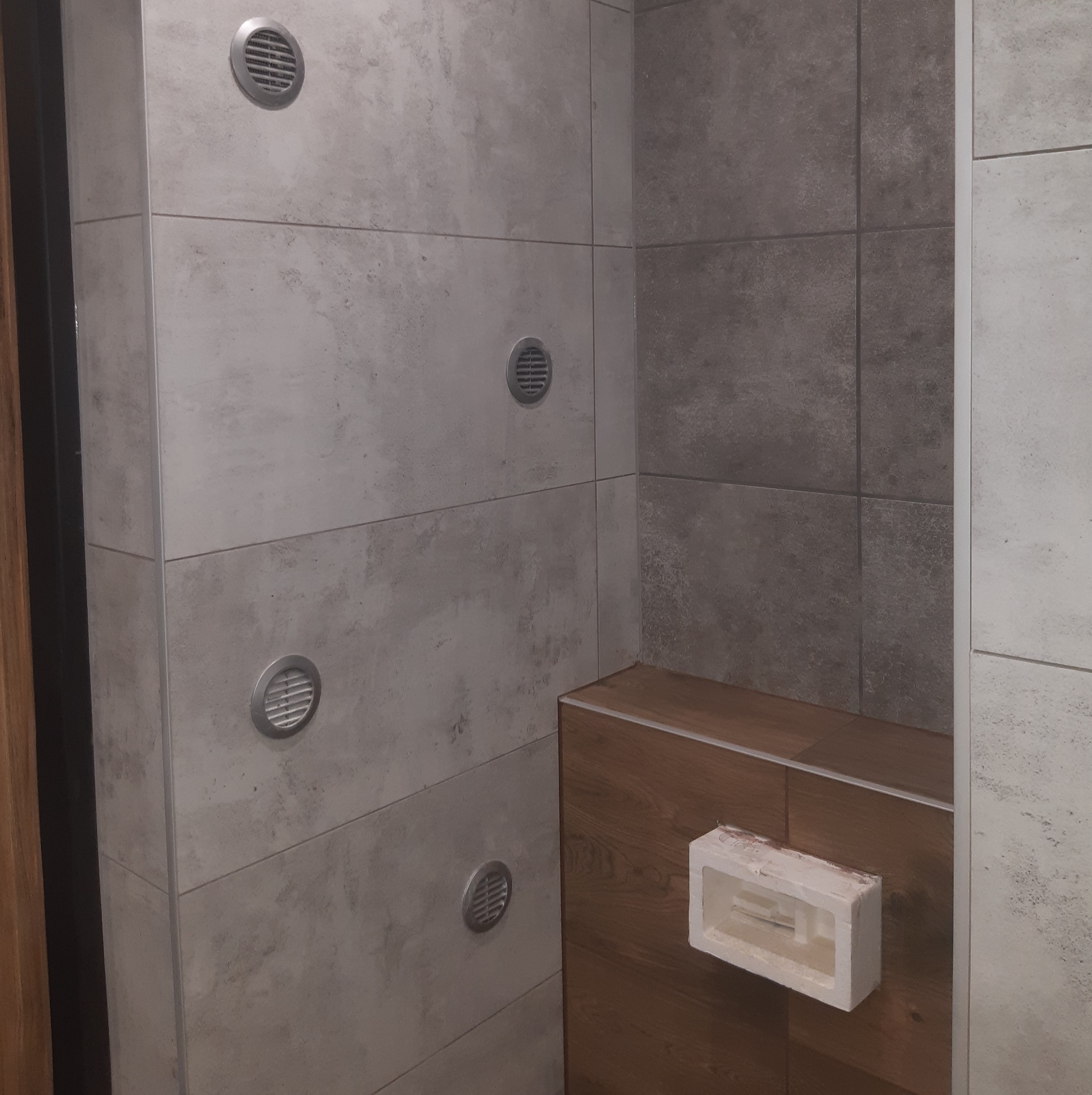 Wykończona łazienka z szarymi płytkami imitującymi beton, drewnianym obudowaniem wanny i kratkami wentylacyjnymi w ścianie, widok na narożnik pomieszczenia.