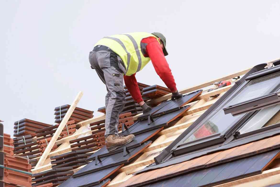 Dekarz w odblaskowej kamizelce montuje ciemną dachówkę na dachu z oknami połaciowymi, na przygotowanym stelażu z łat i kontrłat, obok ułożone stosy dachówek.