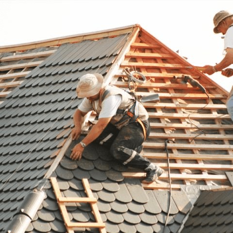 Dekarz w kapeluszu i uprzęży montuje łuskę na dachu, widoczne drewniane łaty i narzędzia, drugi dekarz w tle mierzy odległość.
