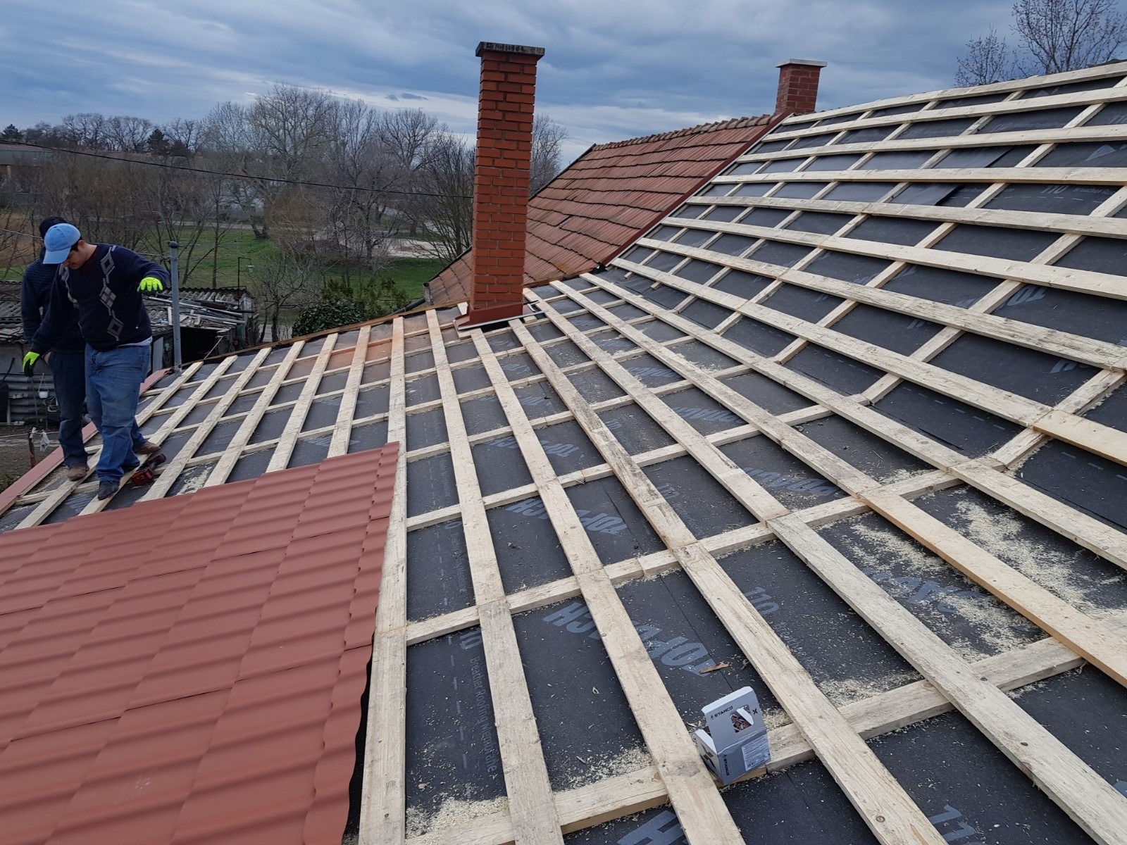 Montaż łat na membranie dachowej z widocznymi fragmentami czerwonej dachówki i kominem w tle, dwóch dekarzy w roboczych ubraniach przy pracy.