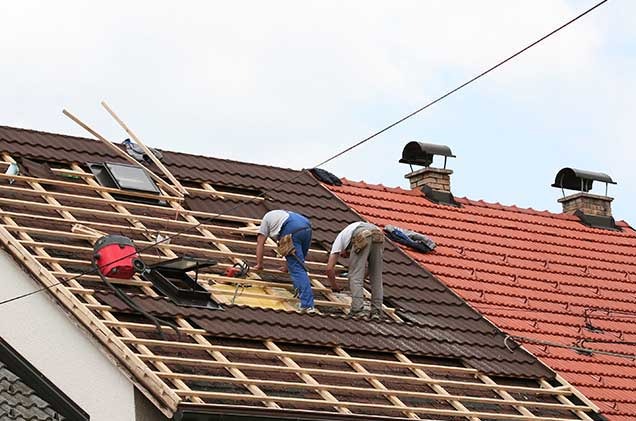 Dwóch dekarzy pracuje na dachu domu, montując drewnianą konstrukcję pod pokrycie z brązowej blachodachówki, widoczny fragment dachu pokryty czerwoną dachówką oraz kominy.