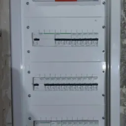 Rozdzielnica elektryczna Schneider z zabezpieczeniami, zamontowana w ramie, widoczna na tle surowej ściany. Kompletny układ z wyłącznikami i ogranicznikami przepięć.