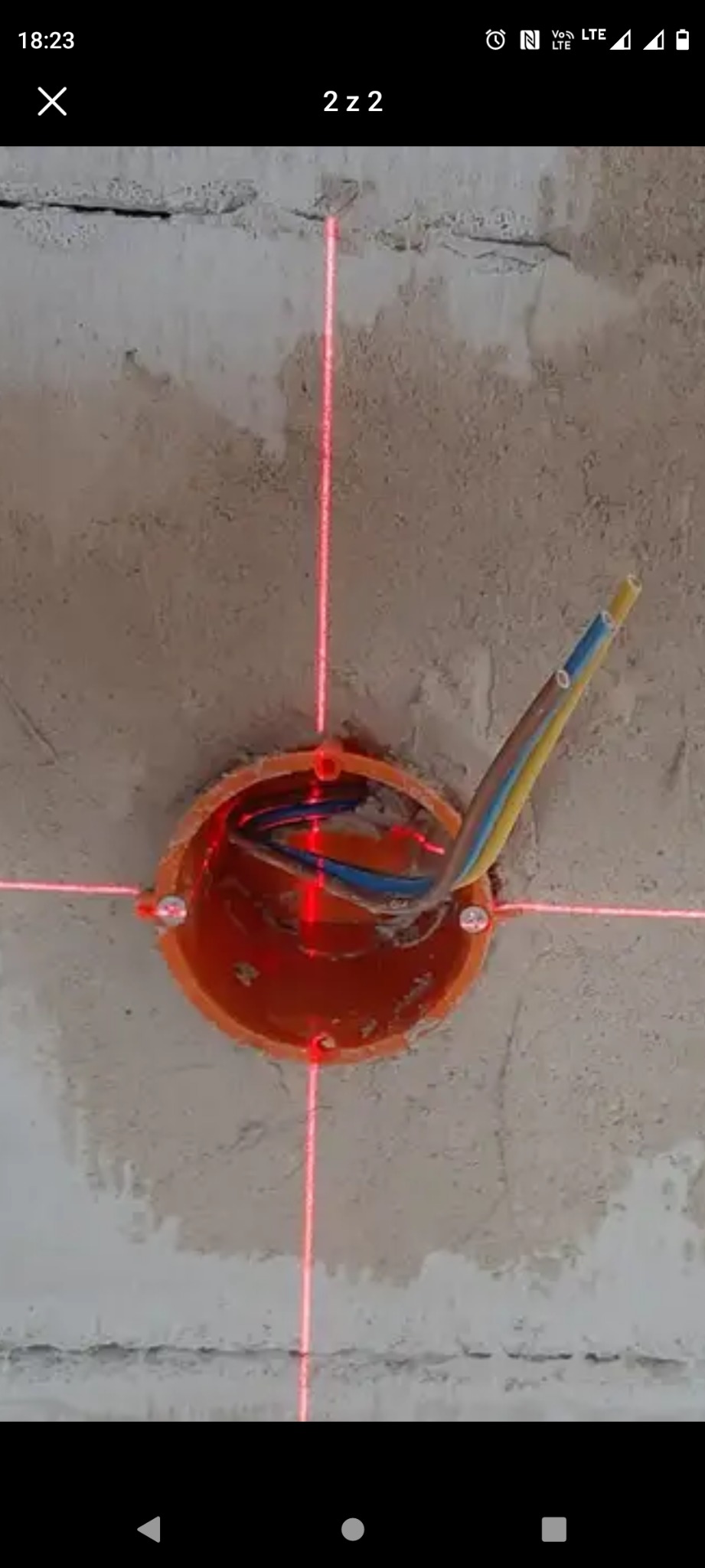 Pomarańczowa puszka elektryczna w surowym suficie, z widocznymi przewodami i liniami lasera krzyżowymi, wyznaczającymi punkty montażowe.