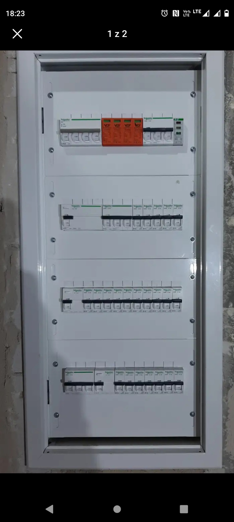 Rozdzielnica elektryczna Schneider z zabezpieczeniami, zamontowana w ramie, widoczna na tle surowej ściany. Kompletny układ z wyłącznikami i ogranicznikami przepięć.