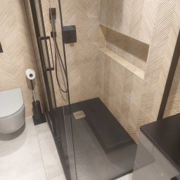 ANDRII KOROL - Elegancka kabina prysznicowa z czarnymi profilami i deszczownicą, wykończona płytkami w jodełkę. Widoczna toaleta i szafka łazienkowa. Nowoczesny design.