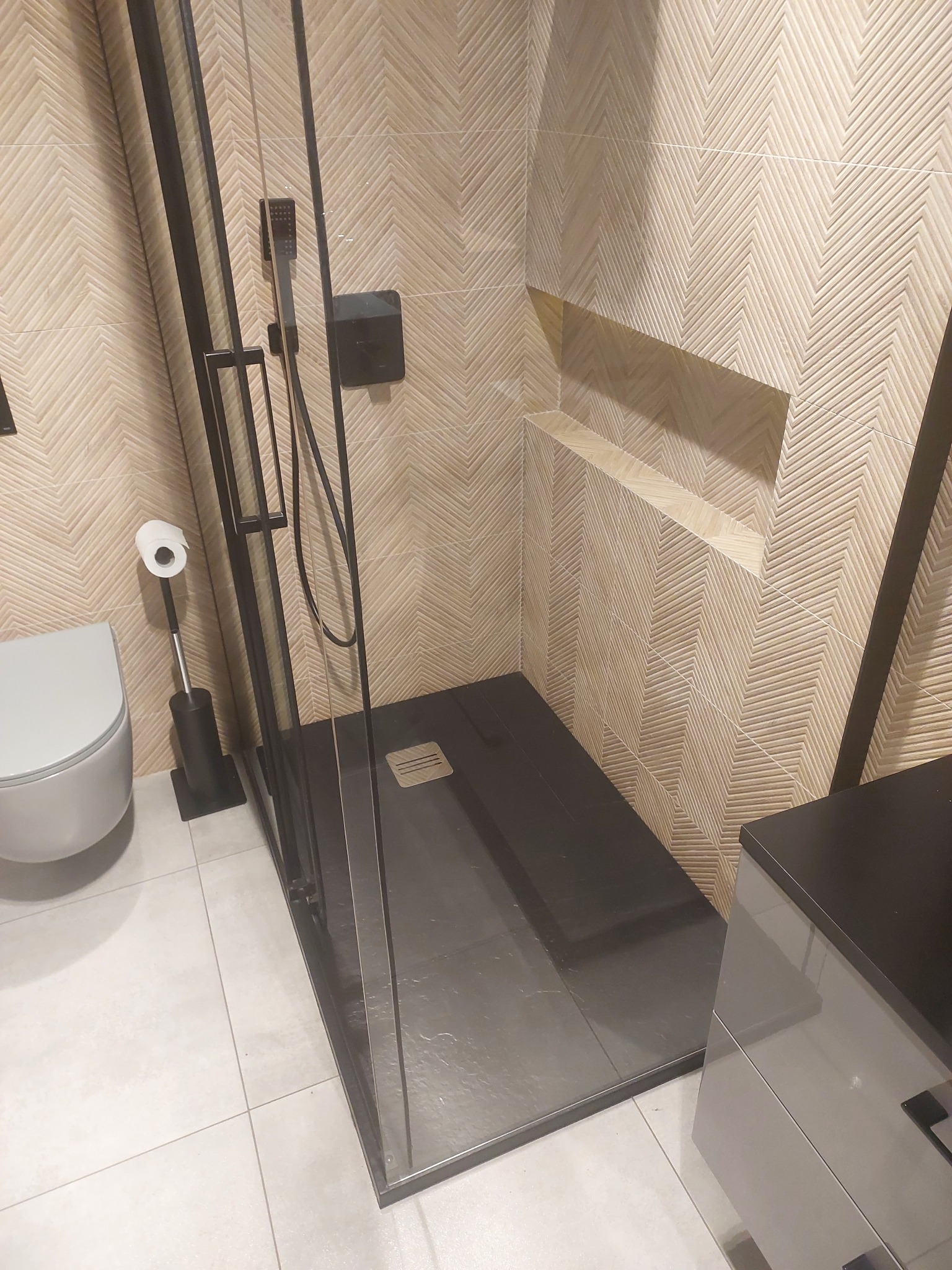 Elegancka kabina prysznicowa z czarnymi profilami i deszczownicą, wykończona płytkami w jodełkę. Widoczna toaleta i szafka łazienkowa. Nowoczesny design.